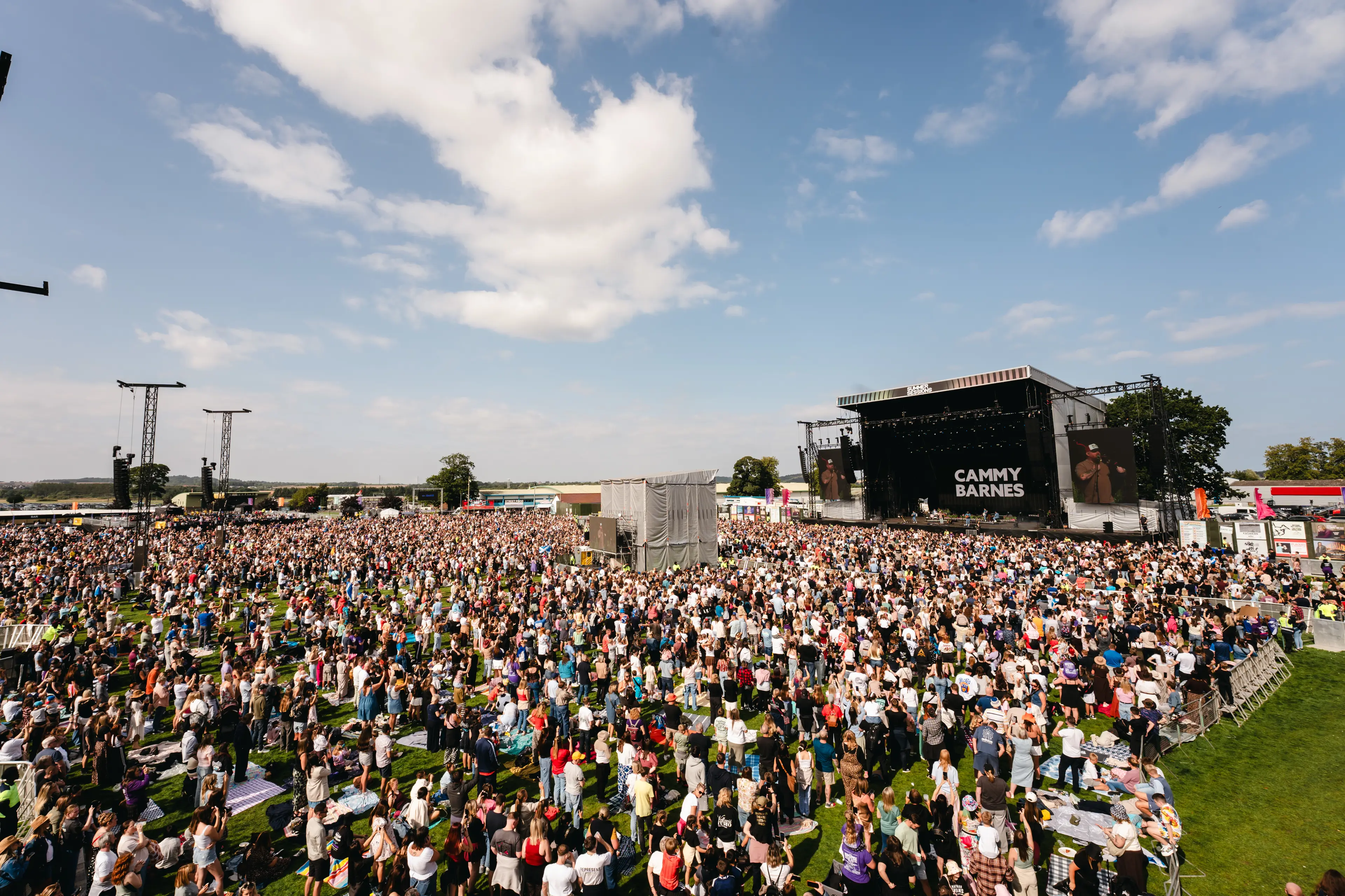 Wide view of a large outdoor crowd watching Cammy Barnes perform on the main stage under a blue sky.
