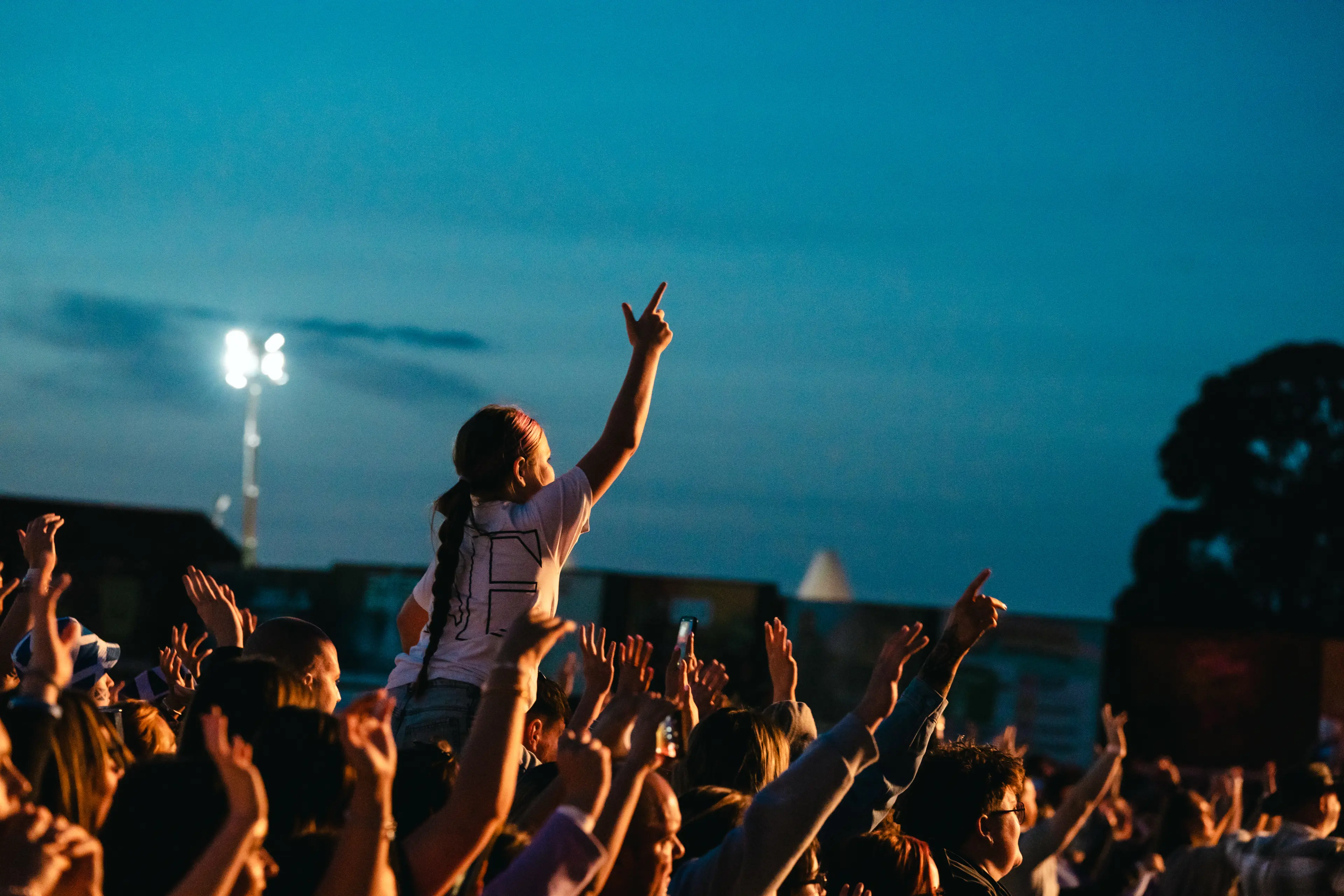 Child sitting on someone’s shoulders raises an arm above a cheering festival crowd at dusk.