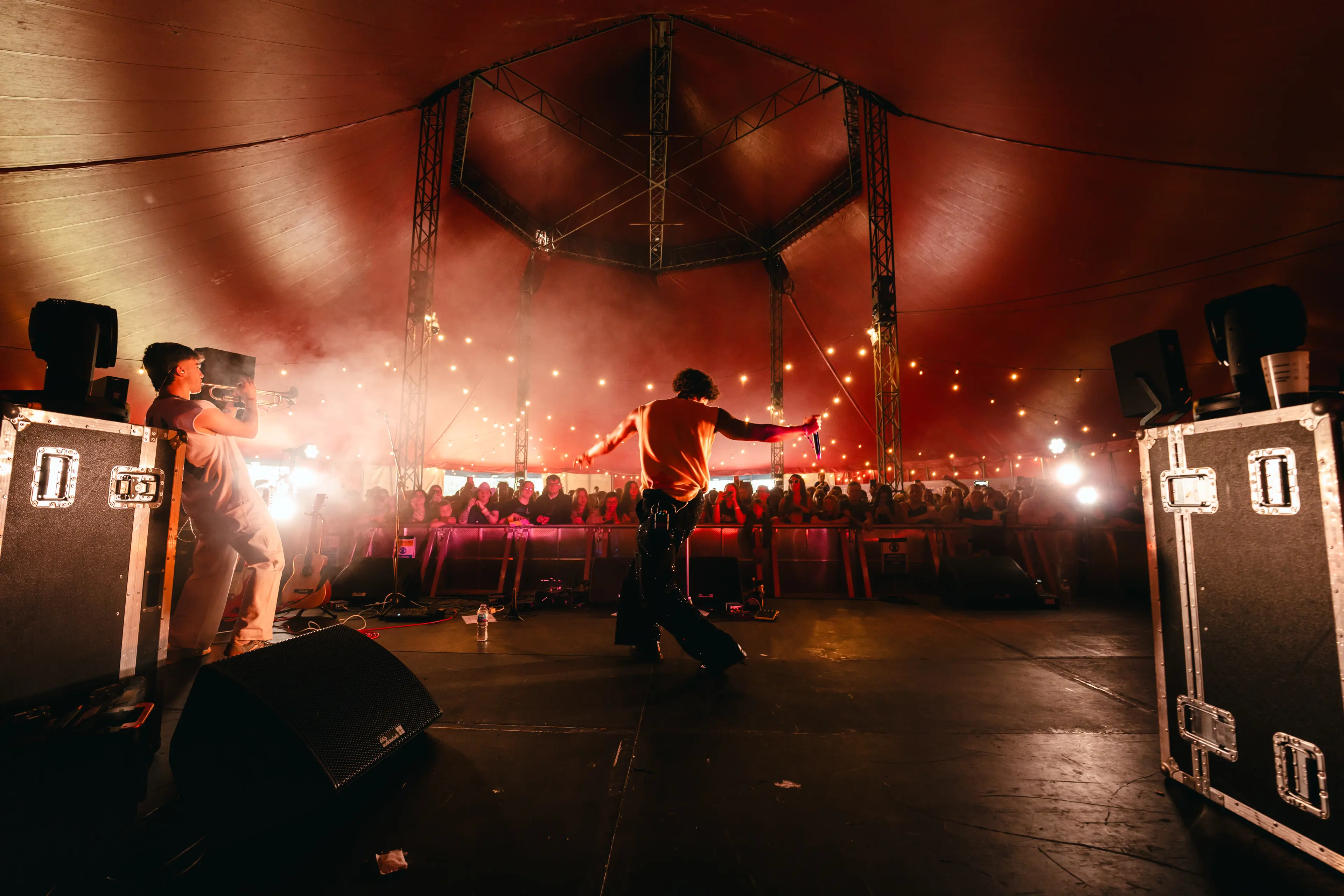 Two performers entertain a crowd from the stage of a warmly lit red festival tent.