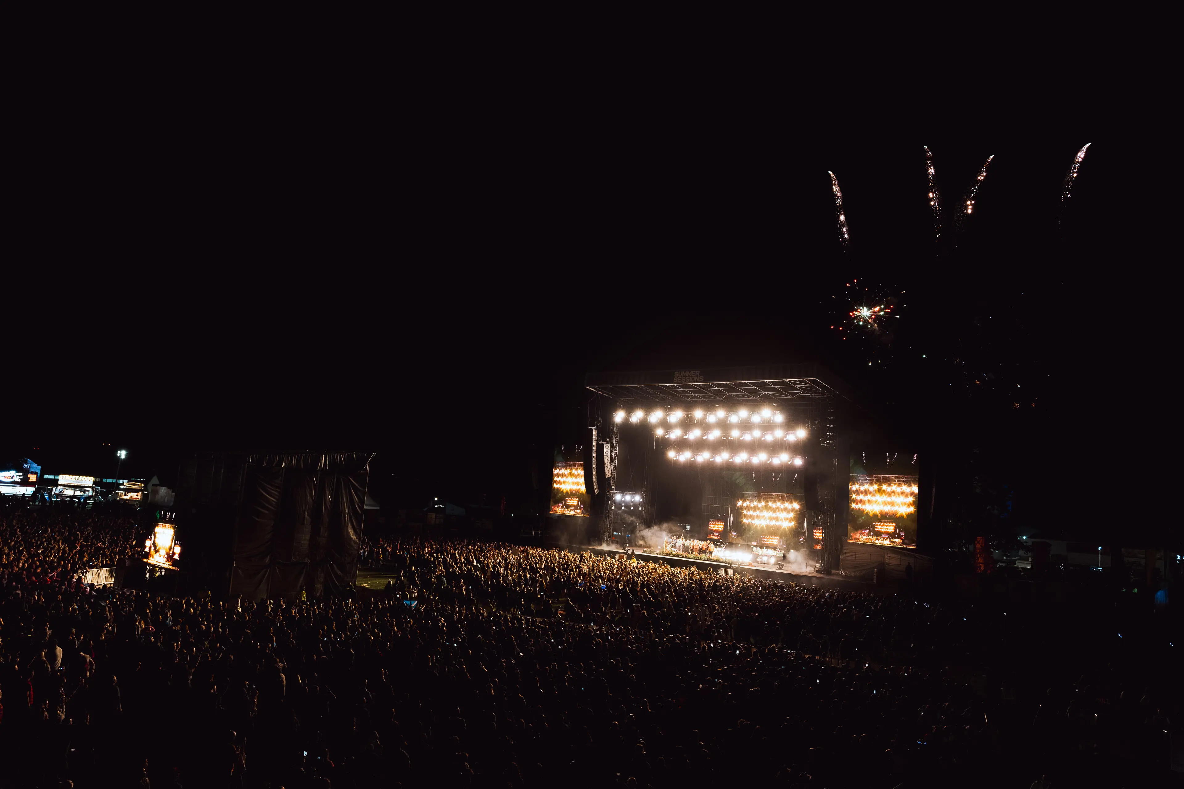 Fireworks burst above a brightly lit festival main stage and packed nighttime audience.