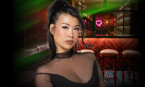 JS im halben Smash Oberteil, im Hintergrund ein pinkes Neonherz und drei Barhocker