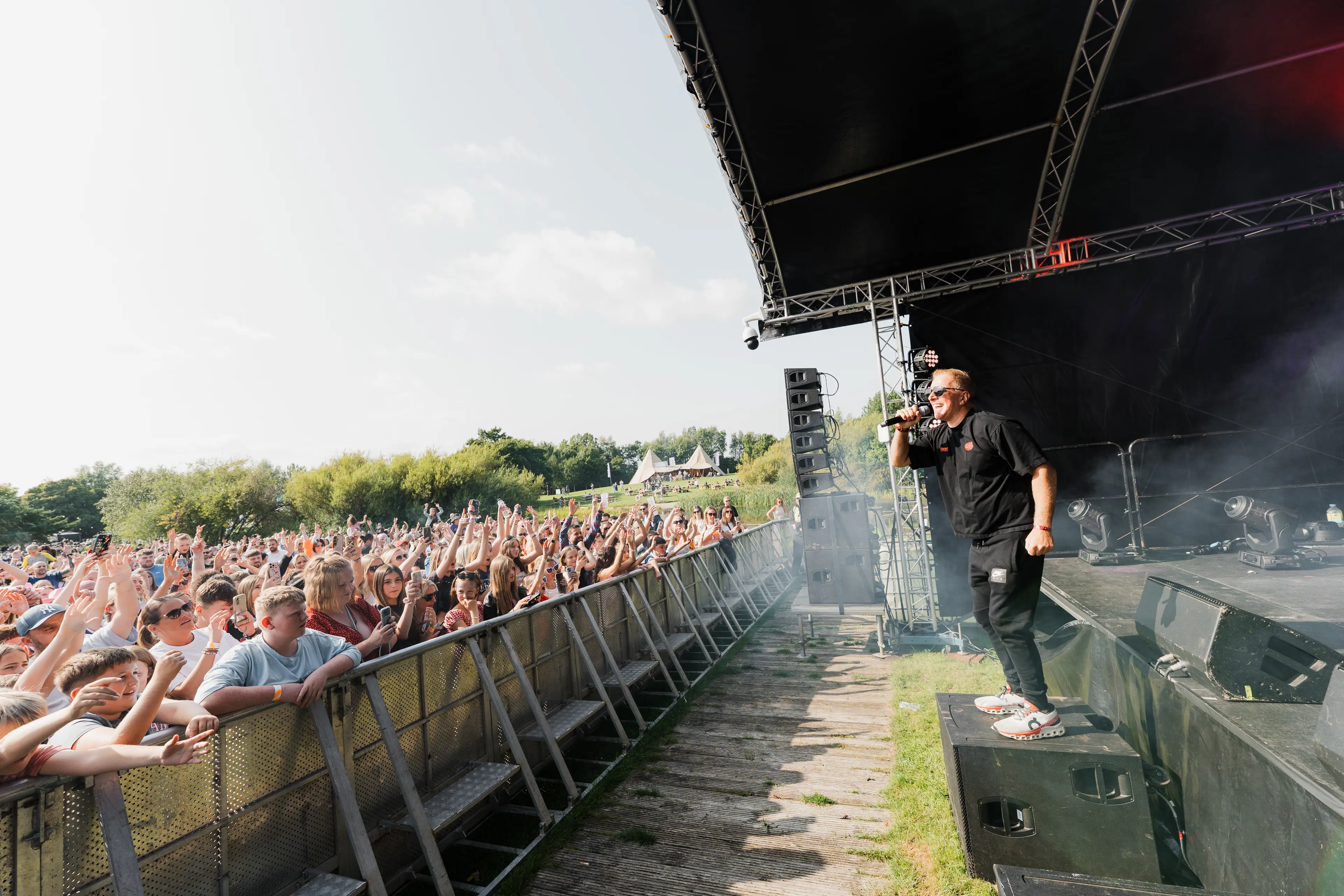 Performer in black addresses a cheering daytime festival crowd from the edge of an outdoor stage.