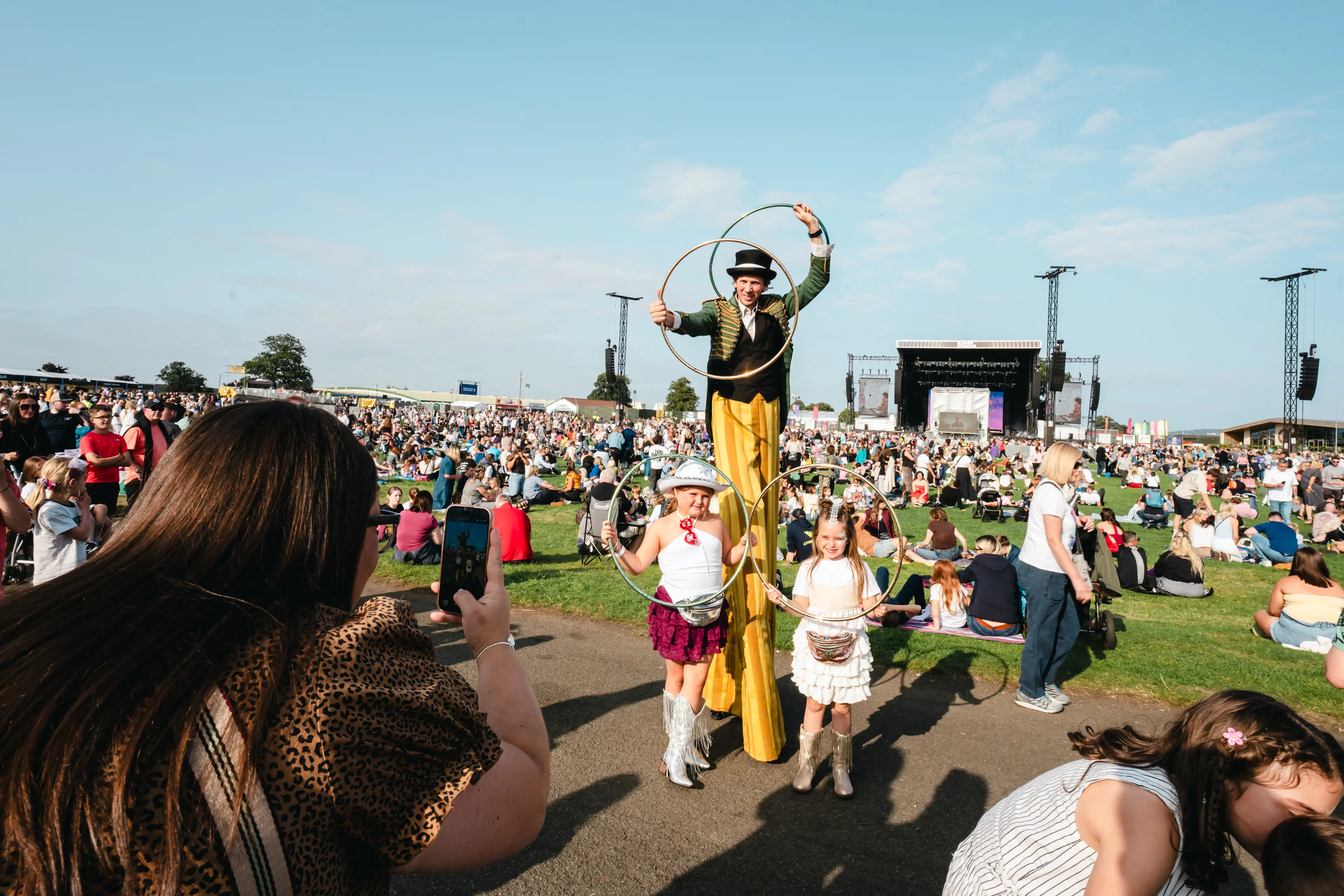 Stilt walker in a green jacket poses with two children holding hoops at a sunny outdoor festival.