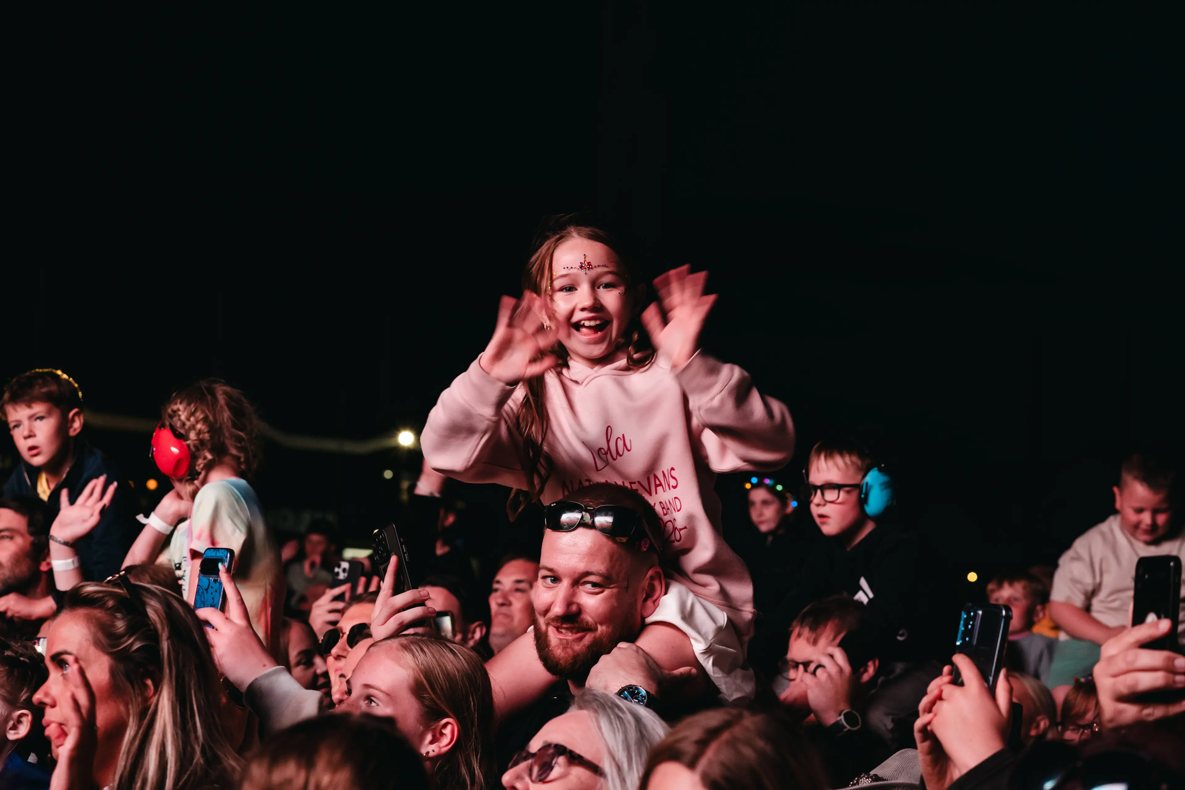 Smiling child on an adult’s shoulders waves above a nighttime festival crowd.