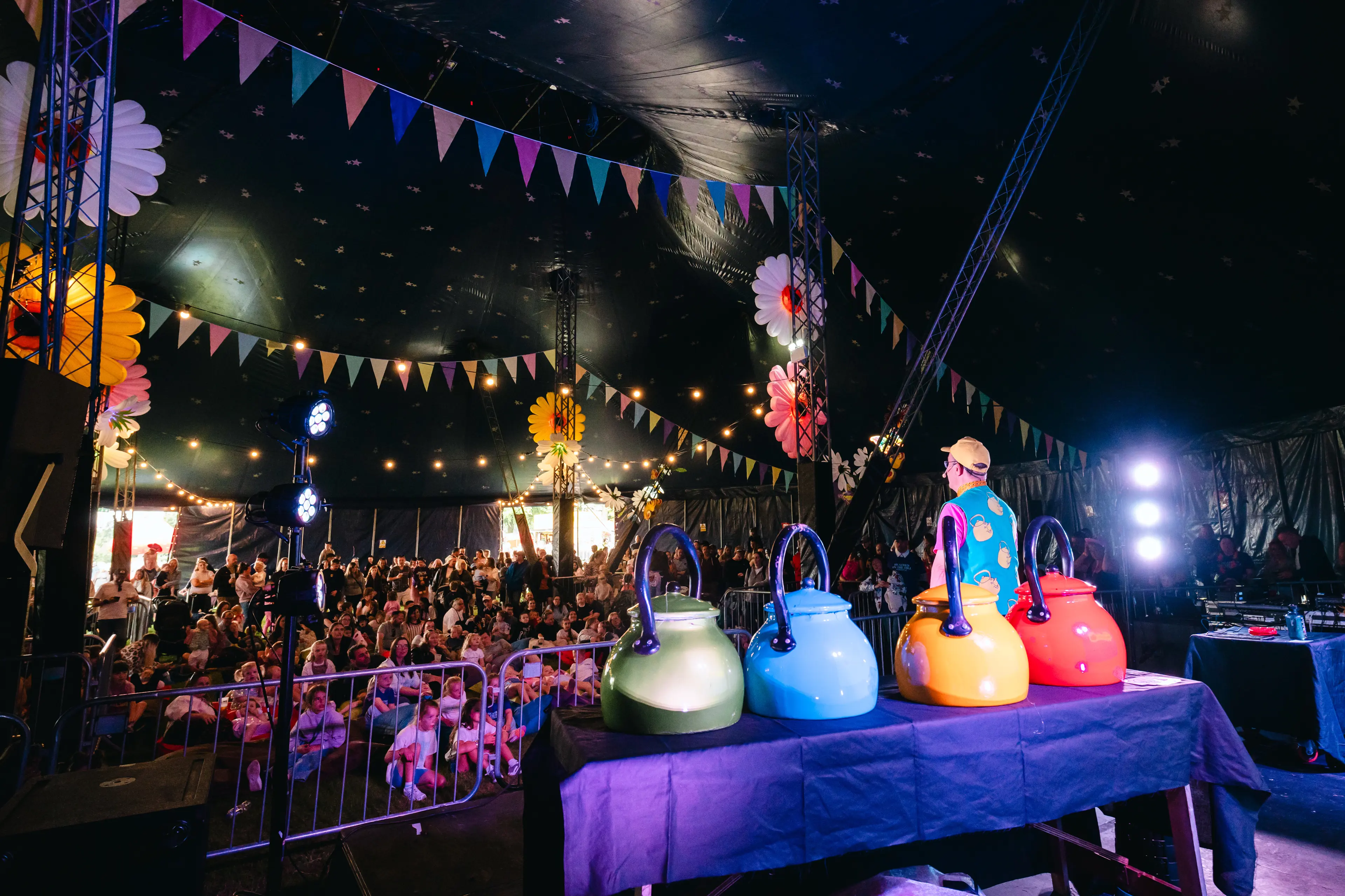 Performer stands beside four oversized colourful kettle props in a bunting-decorated family festival tent.