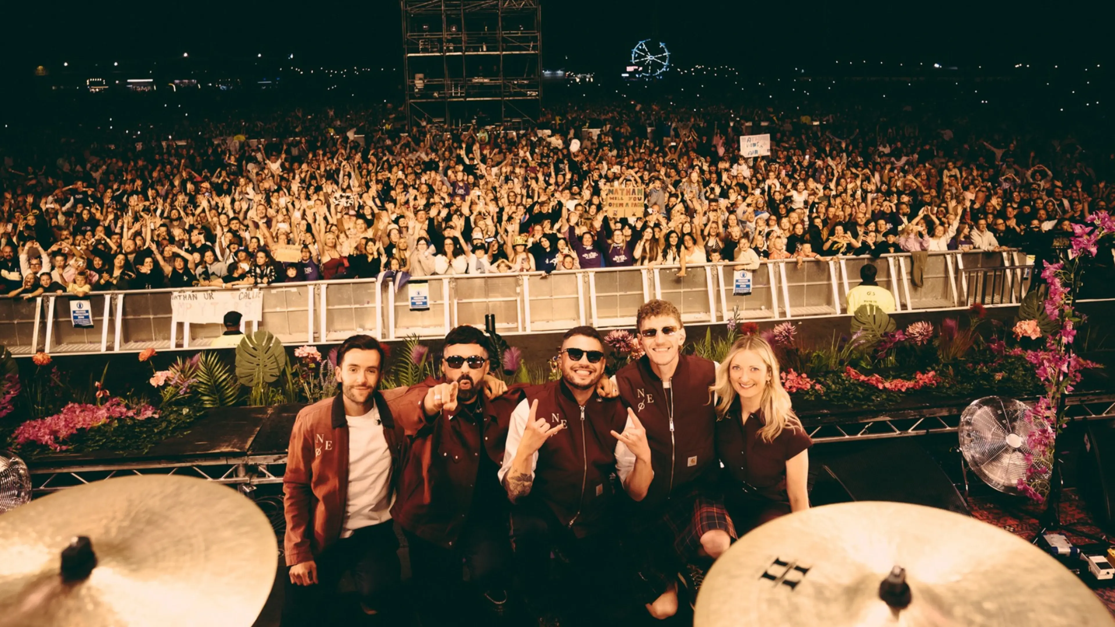 Five performers pose onstage behind a drum kit, with a vast nighttime crowd cheering behind them.