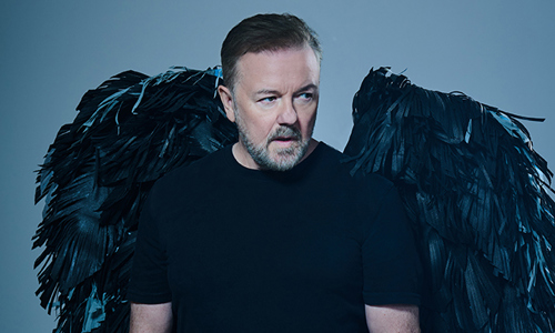 Ricky Gervais im schwarzen T-Shirt und schwarzen Engelsflügeln auf blau/grau meliertem Hintergrund