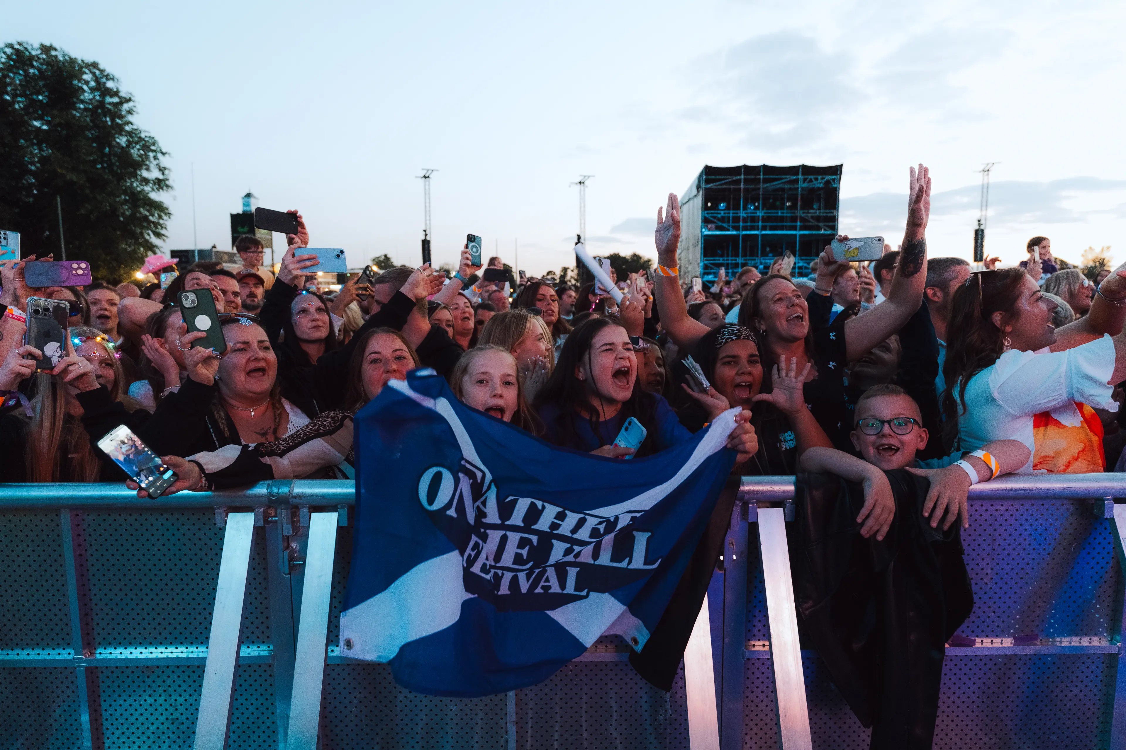 Excited front-row crowd waves phones and an “On the Hill Festival” flag at dusk.