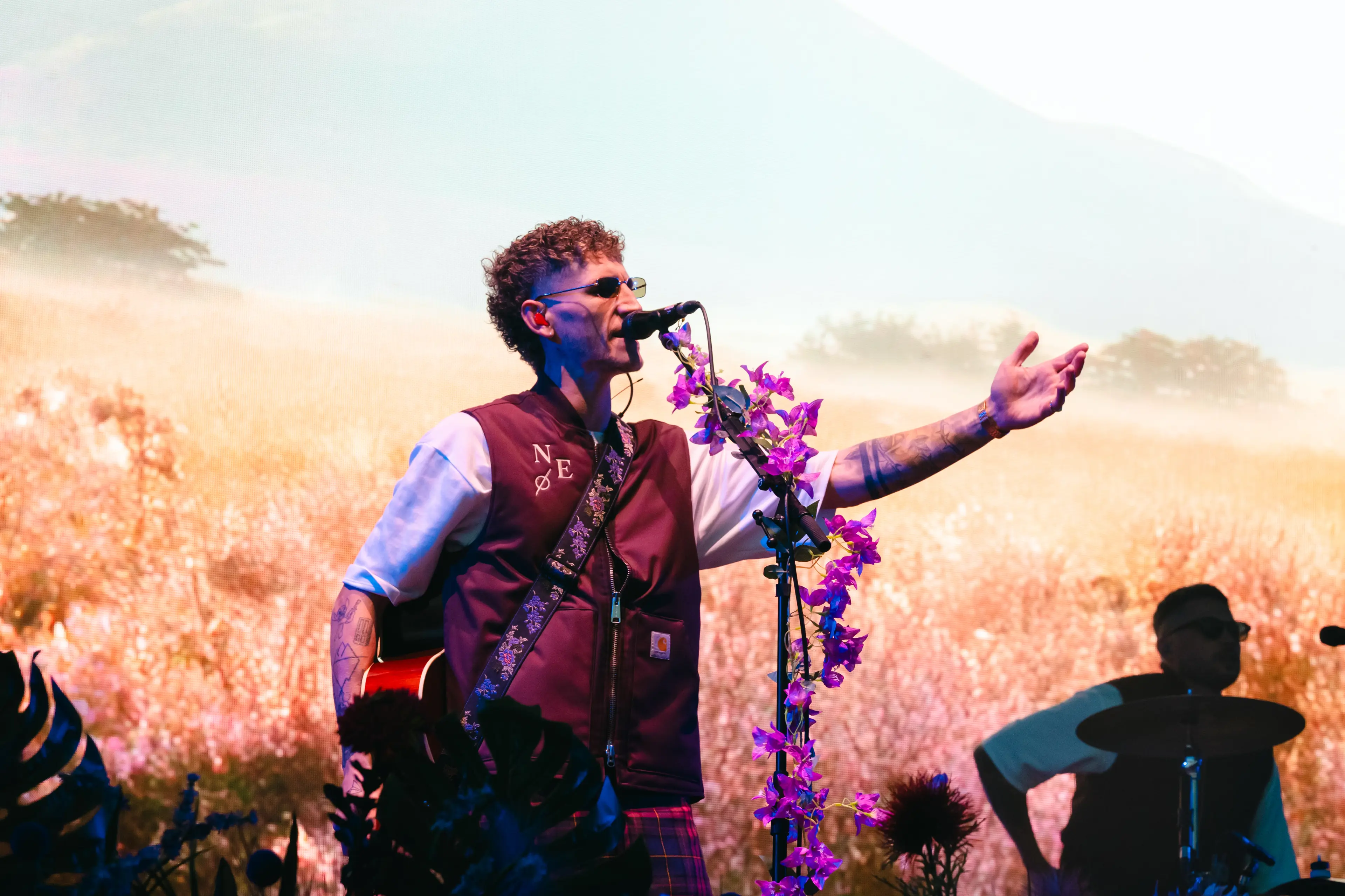 Nathan Evans sings into a flower-decorated microphone onstage, with a drummer behind him.