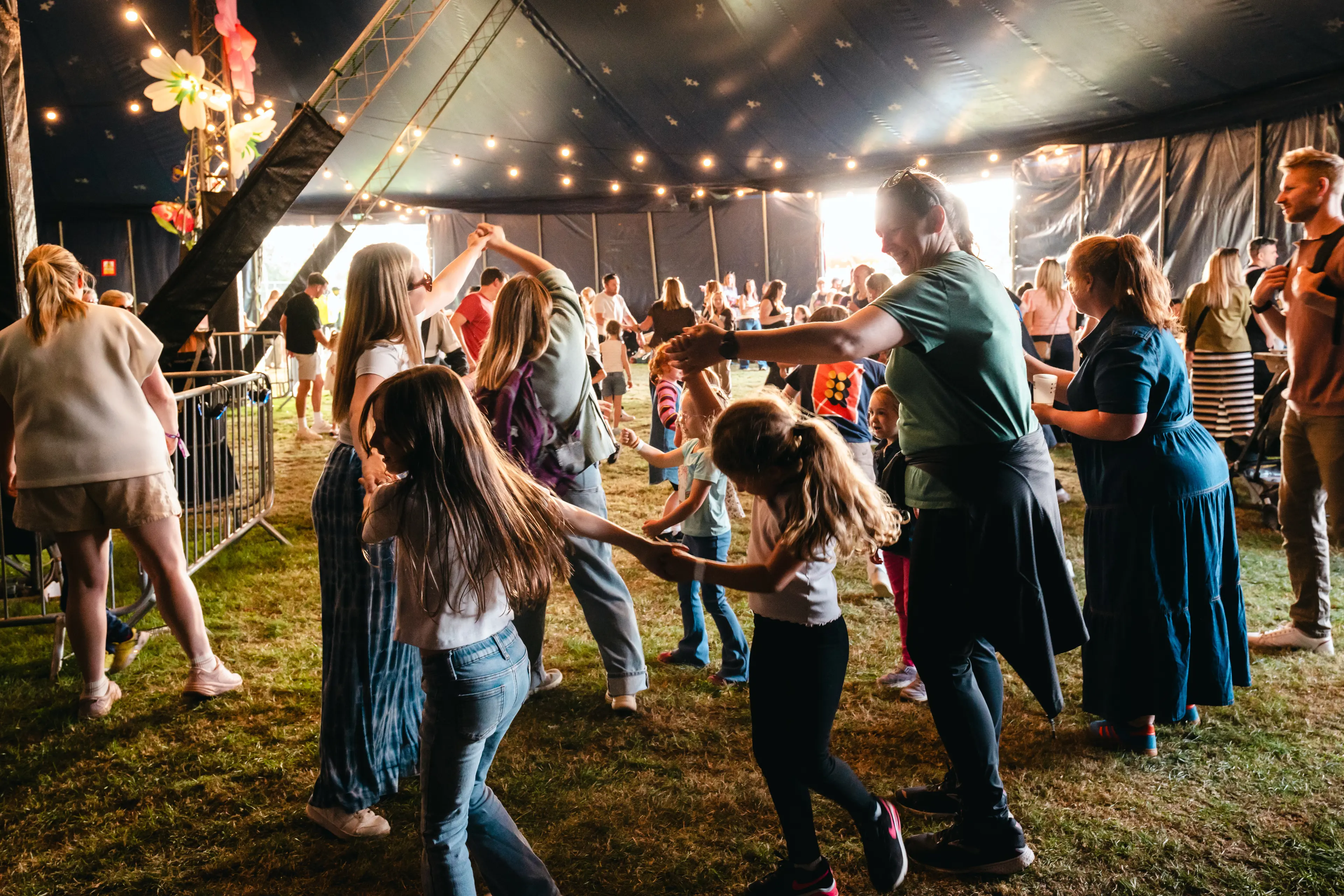 Children and adults join hands and dance a ceilidh inside a decorated festival tent.