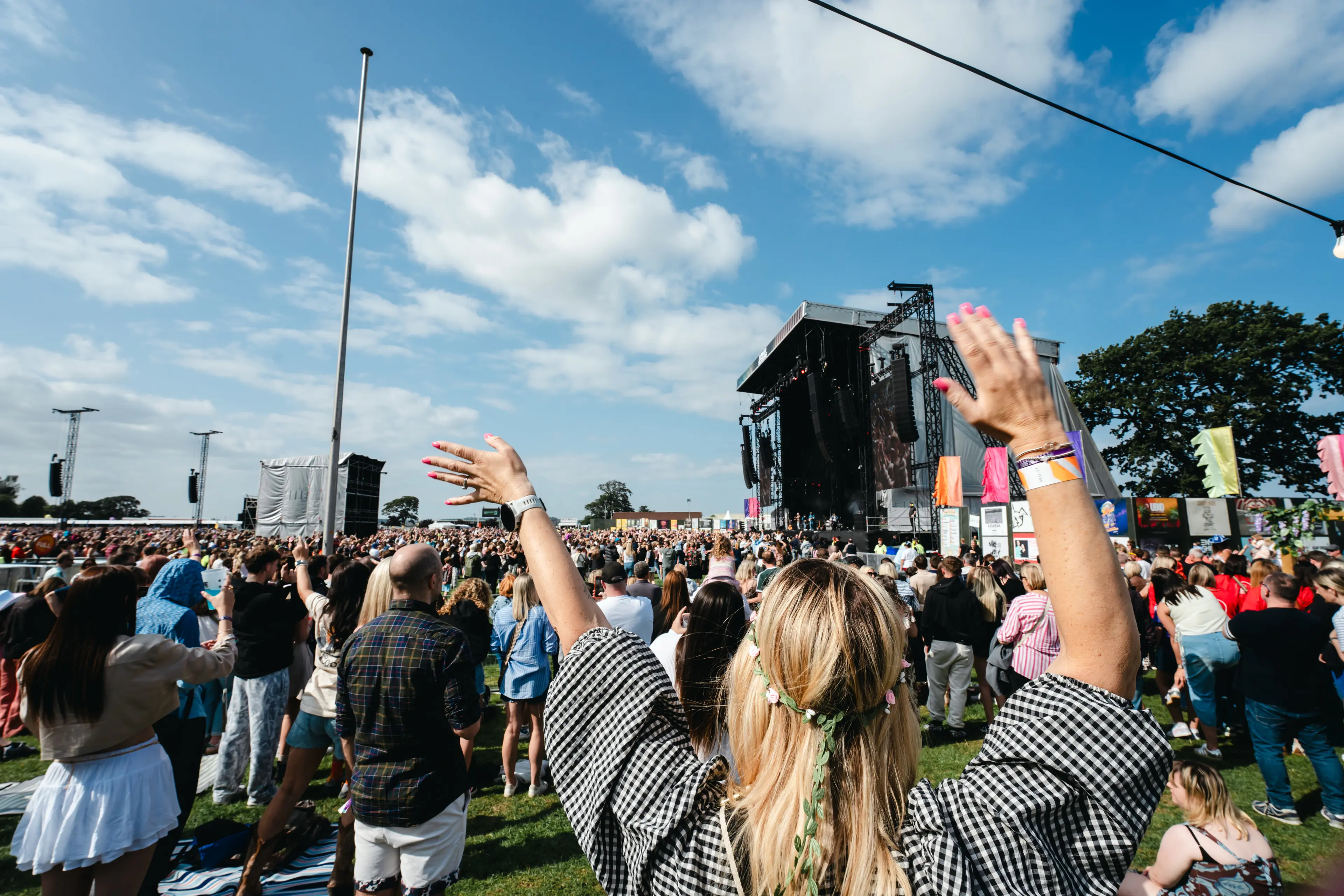 Festivalgoer raises both hands while facing the main stage in a large daytime crowd.