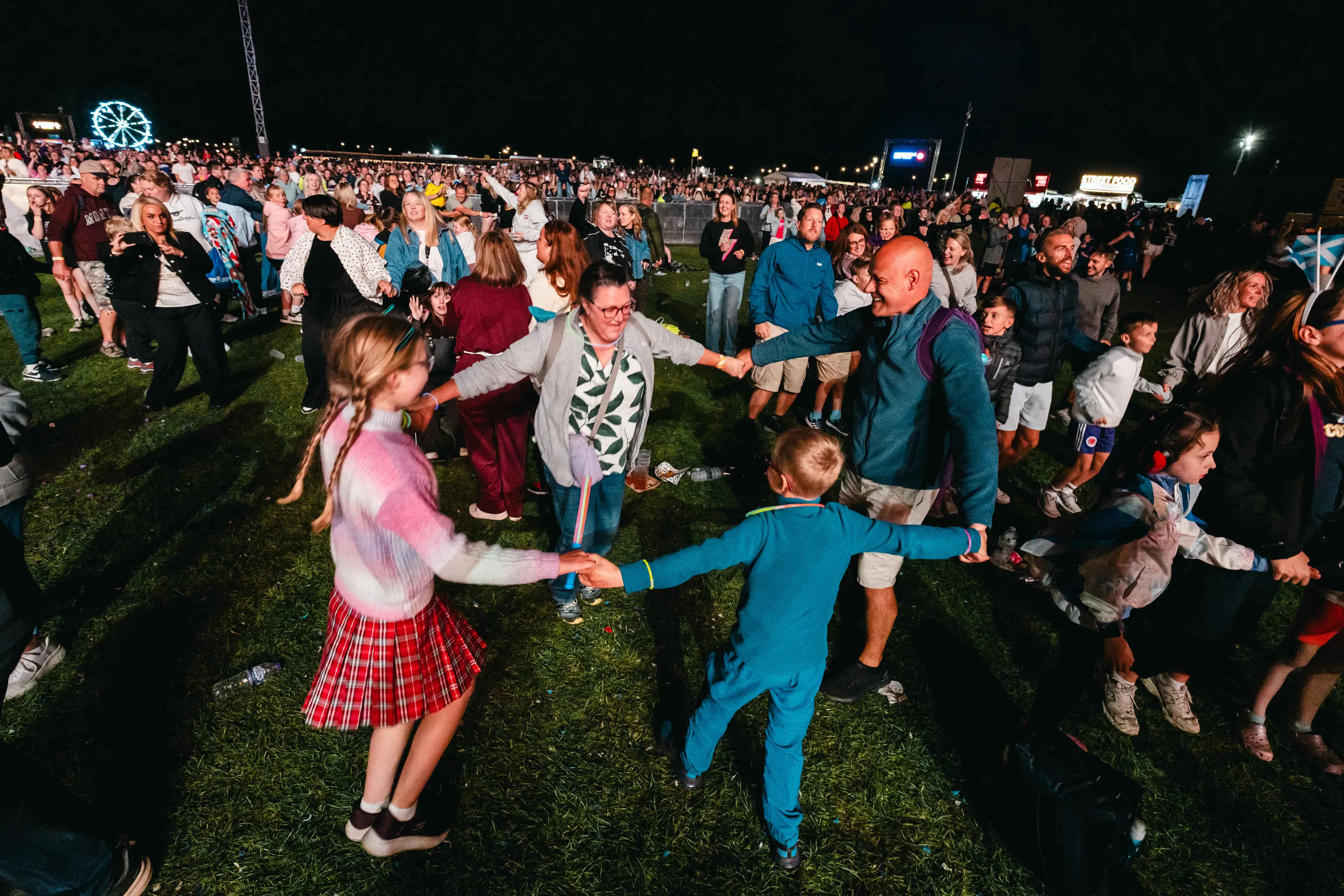 Families and children dance in a circle on the grass at a nighttime festival, with a lit Ferris wheel in the background.